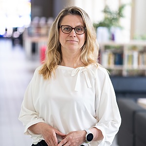 Dr. Laura Daniusevičiūtė-Brazaitė stands in a modern interior with books in the background, wearing a white blouse and holding her hands clasped. She is a senior researcher and associate professor of the project.