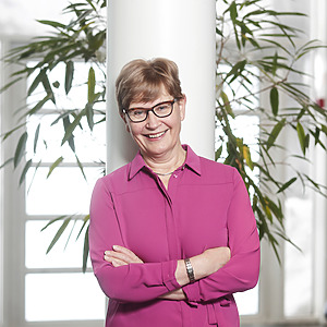 Dr. Anna-Maija L&auml;ms&auml; stands by the window with plants in the background, wearing a pink blouse and smiling. She is the co-leader of the project from the University of Jyv&auml;skyl&auml; in Finland and a professor emerita.