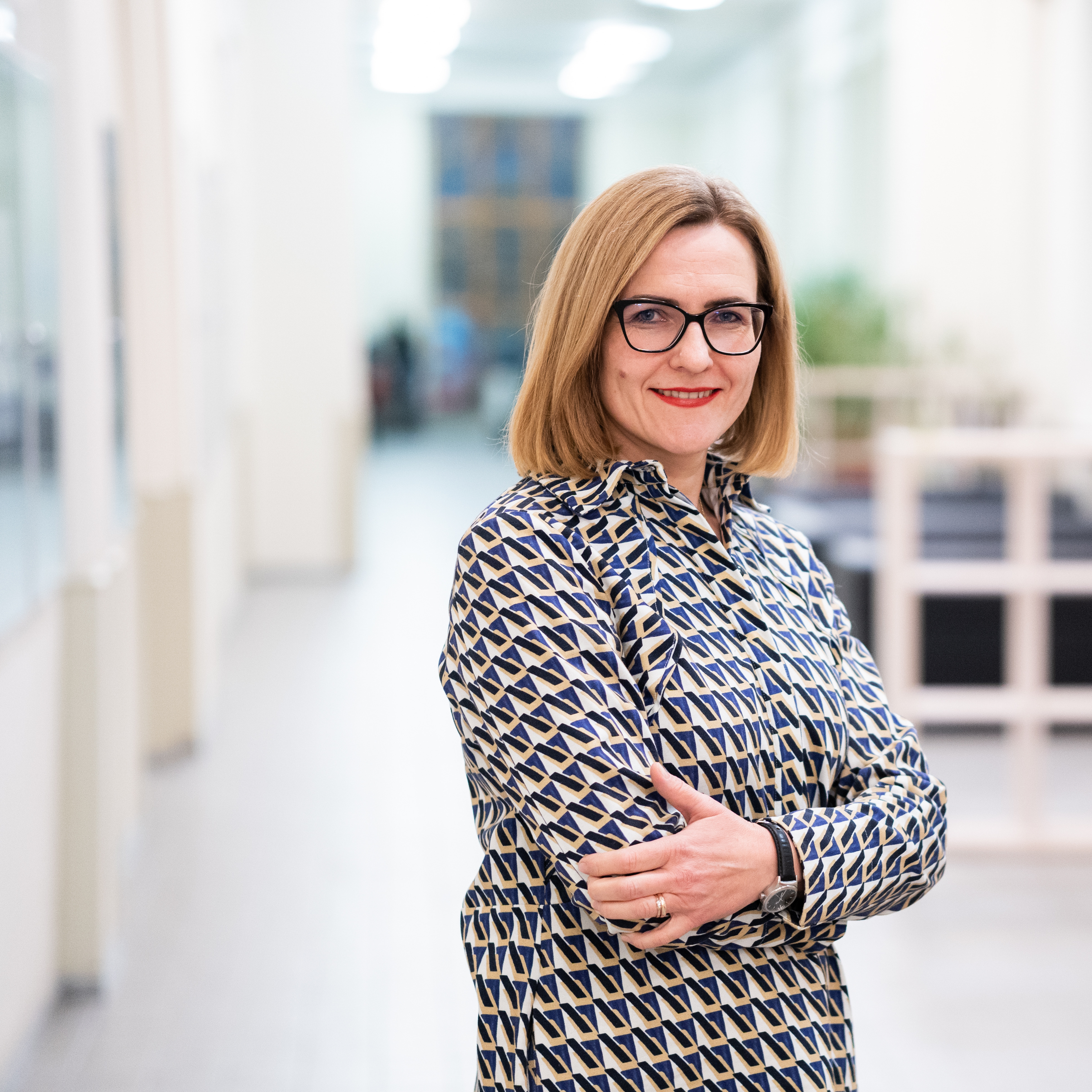 Dr. Raminta Pučėtaitė stovi šviesiame koridoriuje, vilkinti raštuotą suknelę ir besišypsanti, sukryžiavusi rankas. Ji yra PULSE projekto vadovė ir profesorė.