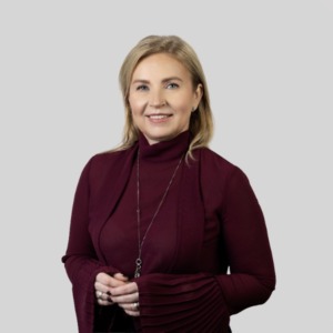 Jolanta Vaičiūnienė stands in a bright background, wearing a red-purple blouse with pleated sleeves, her arms folded in front of her. She looks at the camera and smiles.