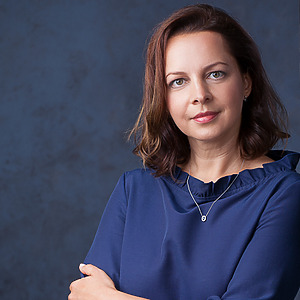 Jūratė Budrienė poses in front of a blue background, wearing a blue blouse and a pendant. She is a junior researcher and doctoral student in the PULSE project.