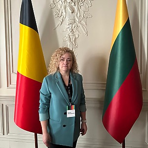 Dr. Donata Jovarauskienė stands between the flags of Belgium and Lithuania, wearing a blue jacket and a name tag. She is the project implementer and holds a PhD in social sciences.