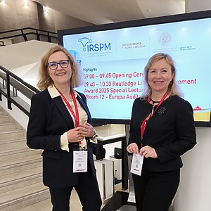 Raminta Pučėtaitė and Jolanta Vaičiūnienė, wearing black jackets and smiling, stand in front of the IRSPM conference display board.
