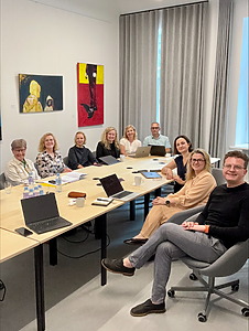 In a bright office, surrounded by colorful paintings, nine members of the PULSE project team (three on the right and six on the left) are seated around a long table conducting research on petty corruption in the health sector.